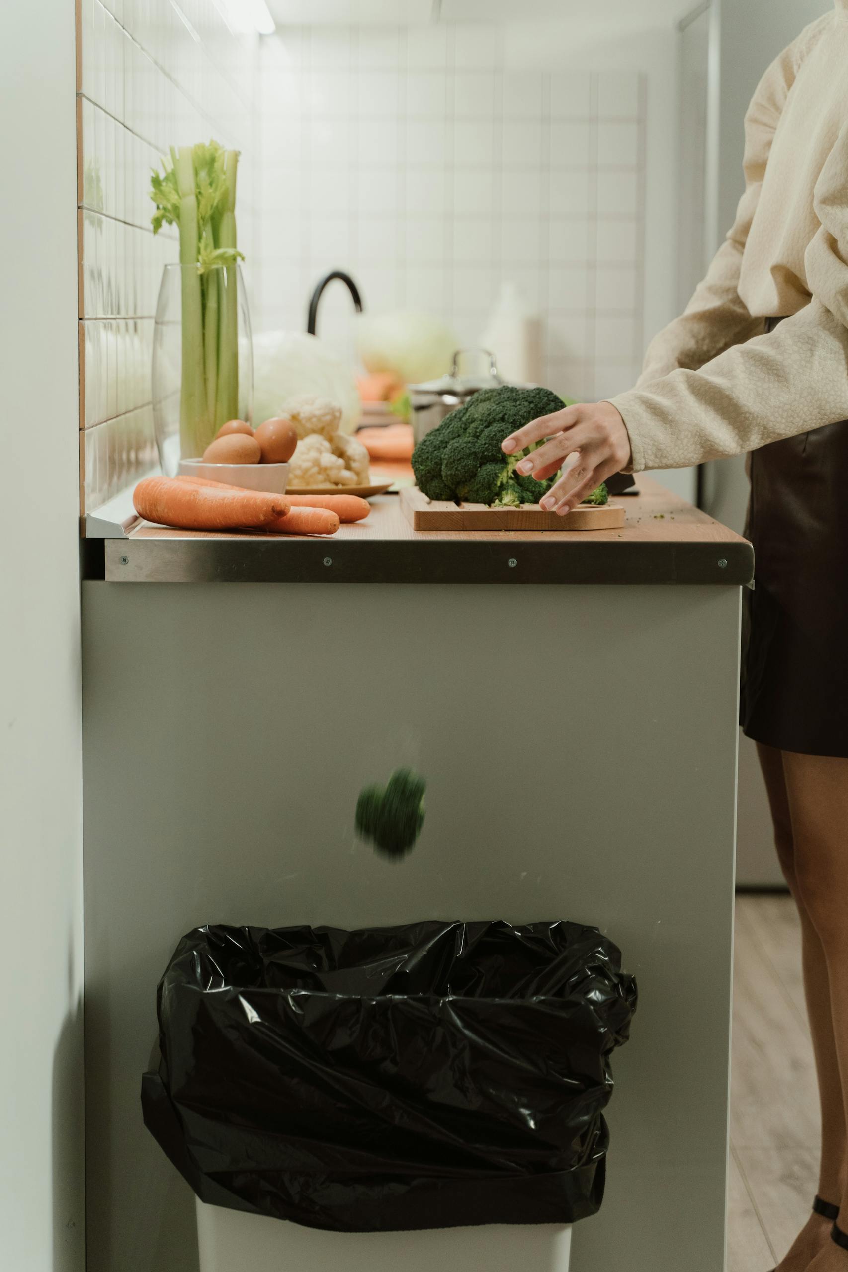 Person discarding broccoli in a stylish kitchen with fresh vegetables on the counter.